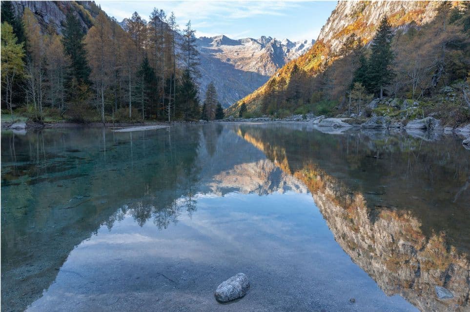 Un lago calmo e limpido riflette una valle montana con alberi autunnali e lontane cime innevate sotto un cielo azzurro.