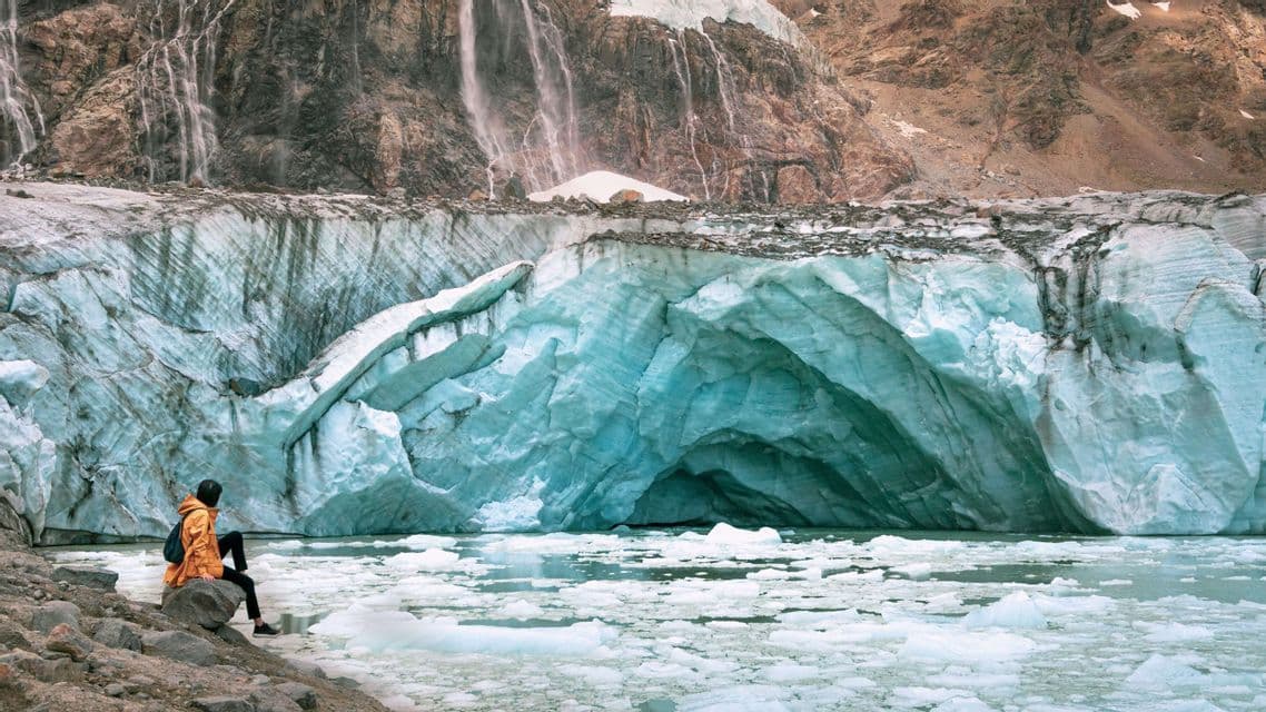 Una persona con una giacca arancione siede su una riva rocciosa, guardando un grande ghiacciaio turchese con una grotta di ghiaccio su un lago con ghiaccio galleggiante.