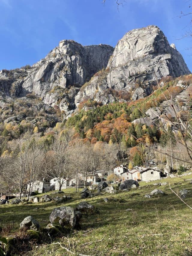 Un piccolo villaggio di case in pietra su un pendio erboso alla base di una massiccia montagna rocciosa con alberi autunnali.