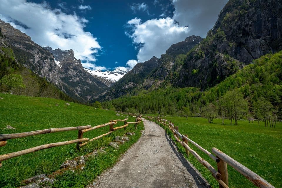 Un sentiero di ghiaia con recinzioni in legno si snoda attraverso una valle verde verso montagne innevate sotto un cielo azzurro nuvoloso.