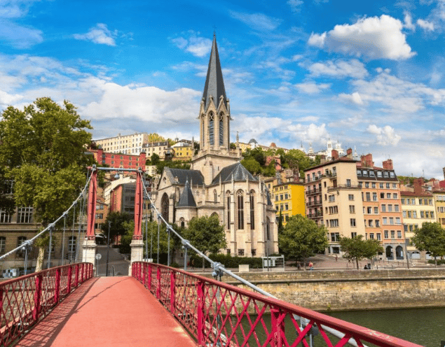 Un ponte pedonale rosso conduce verso una chiesa storica e edifici colorati su una collina sotto un cielo parzialmente nuvoloso.