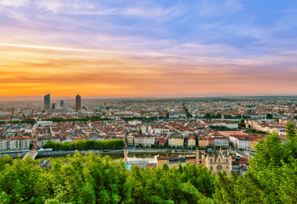 Un ampio panorama urbano con edifici tradizionali e moderni grattacieli si dispiega sotto un tramonto colorato, visto da un punto panoramico elevato sopra alberi verdi.