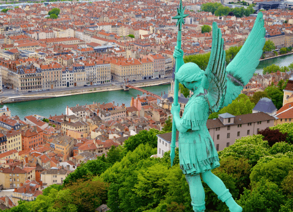 Una statua di angelo con patina verde e un bastone si affaccia su un paesaggio urbano con tetti rossi e un fiume, da un punto panoramico elevato.