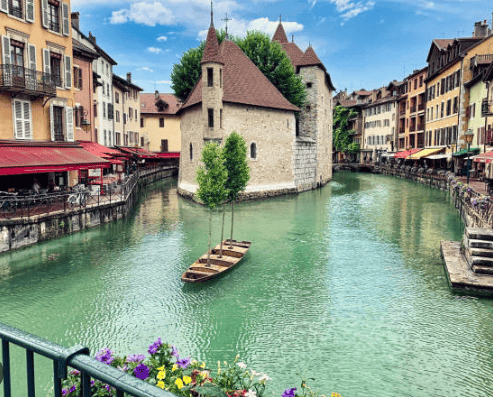 Un edificio medievale in pietra si trova al centro di un canale turchese, affiancato da edifici colorati con ristoranti sotto un cielo blu.