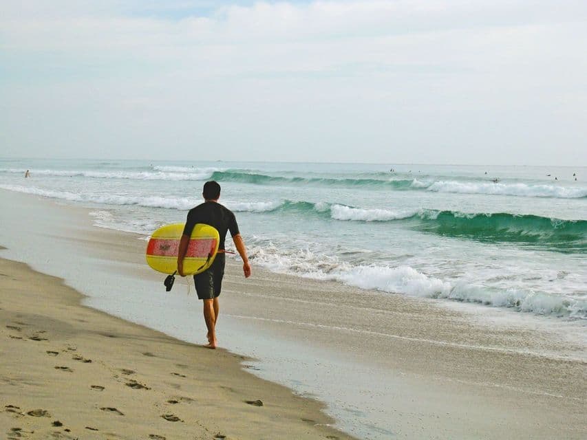 Un surfista in muta cammina lungo una spiaggia sabbiosa, trasportando una tavola da surf gialla verso il mare.