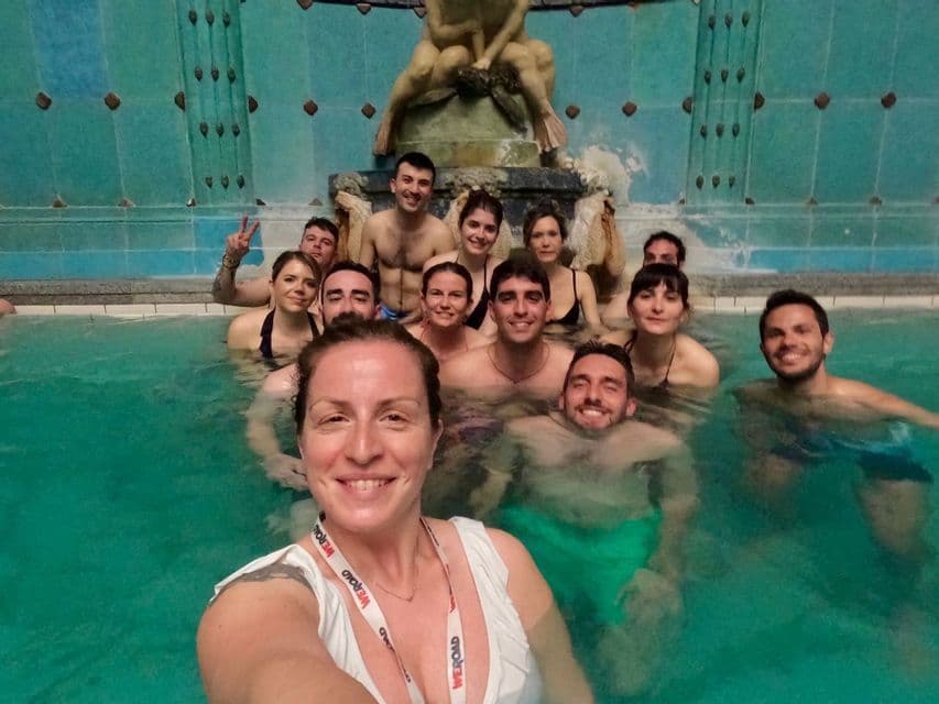 Un grupo de WeRoad se toma una selfie en un baño termal interior, con una pared de azulejos y una escultura al fondo.