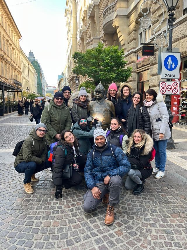 Un viaje en grupo de WeRoad con ropa de invierno posando para una foto grupal con una estatua de bronce en una calle adoquinada de la ciudad.