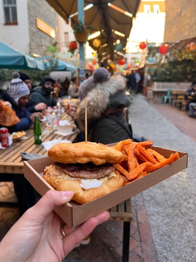 Una mano sostiene una bandeja con una hamburguesa y papas fritas de batata en un mercado de comida al aire libre, con un grupo de viaje de WeRoad comiendo de fondo.