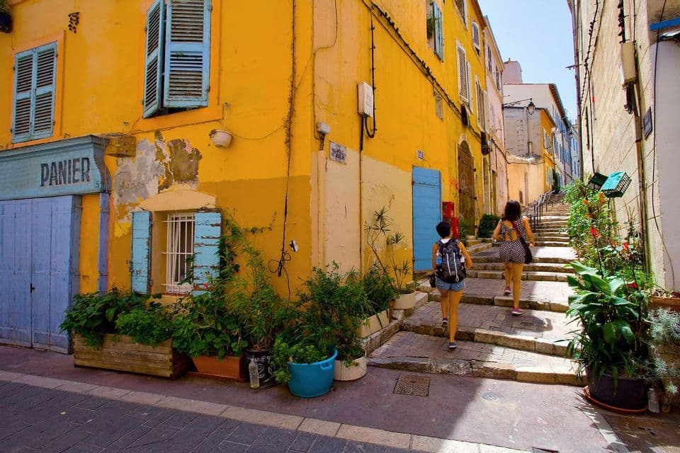 Dos mujeres suben una escalera de piedra en un callejón estrecho flanqueado por edificios amarillos, persianas azules y macetas.