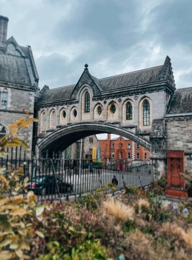 Un puente de piedra con arco de estilo gótico que conecta dos grandes edificios sobre una calle, visto desde detrás de una valla y un jardín.
