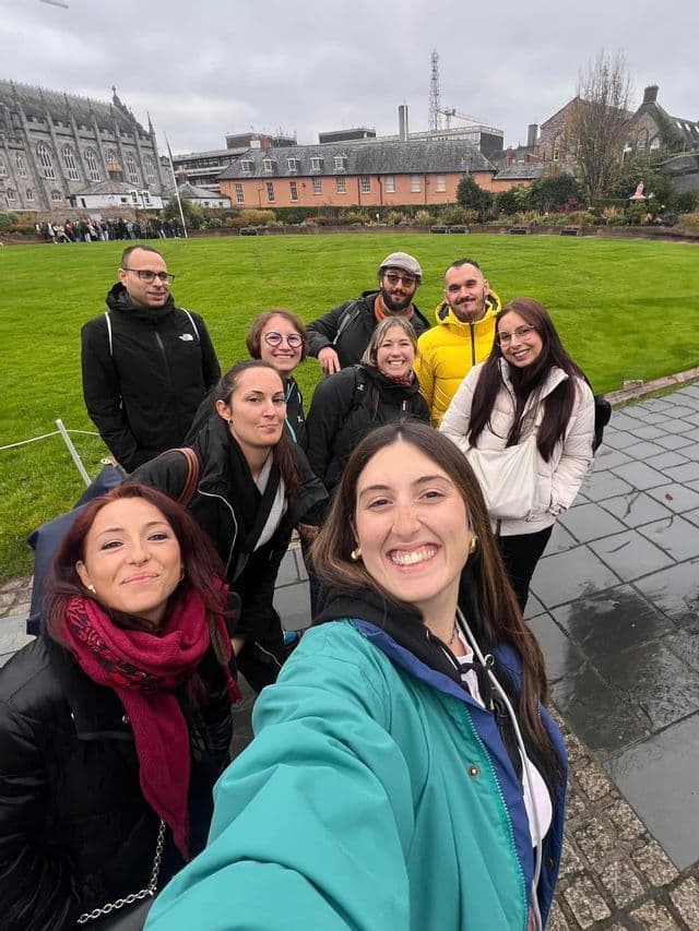 Un grupo de WeRoad se toma una selfie sonriente en un parque verde con edificios históricos de fondo.