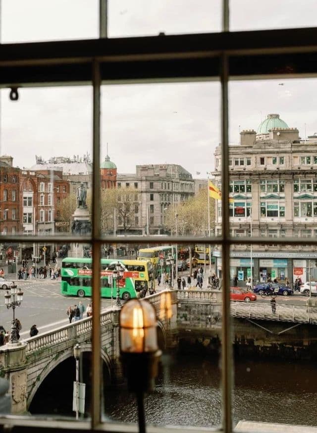 Una bulliciosa calle urbana con un puente de piedra sobre un río y autobuses de dos pisos, vista desde el marco de una ventana.