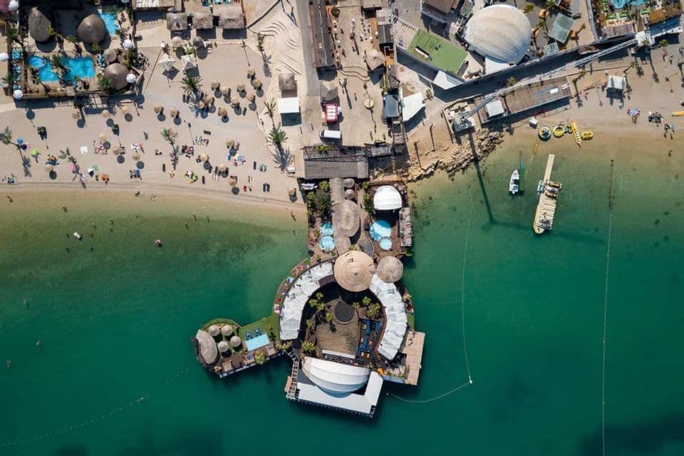 Una vista aerea di un beach club galleggiante con piscine che si estendono da una spiaggia sabbiosa in acqua turchese con barche.