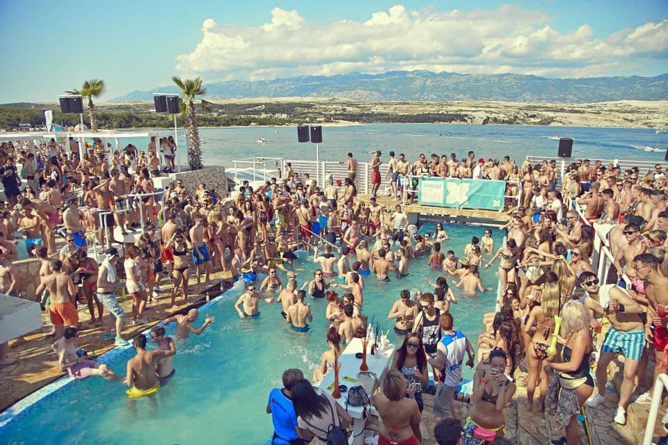 Un folto gruppo WeRoad si gode una festa in piscina di giorno in un club sul mare con montagne sullo sfondo.