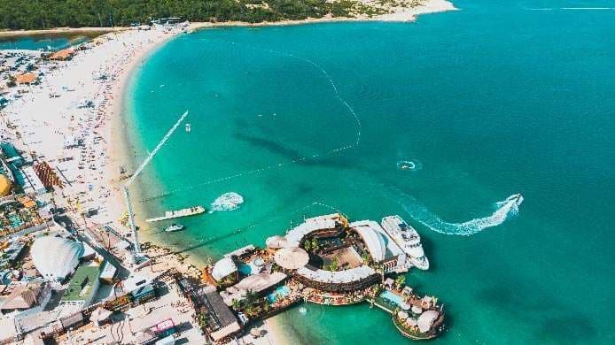 Vista aerea di una spiaggia affollata di sabbia bianca accanto a acqua turchese, con un beach club circolare e barche.