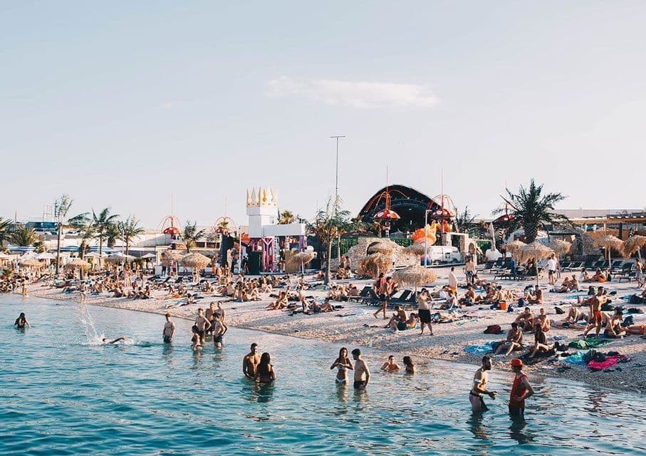 Una grande folla di persone a un festival in spiaggia, con alcuni che nuotano nell'acqua e altri che si rilassano sulla sabbia sotto un cielo sereno.