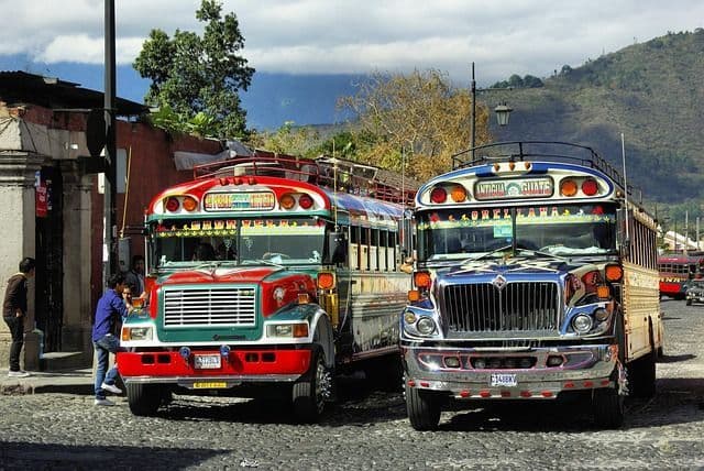 Due autobus colorati, uno rosso e uno blu, sono parcheggiati su una strada di ciottoli con montagne sullo sfondo.