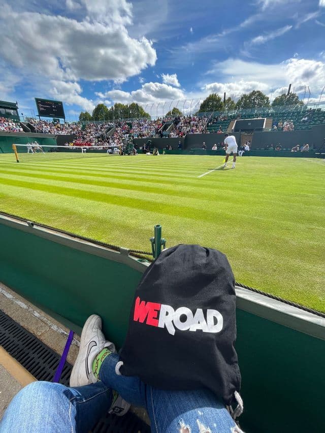 Una vista in prima persona dagli spalti di una partita di tennis su un campo in erba, con una borsa WeRoad nera appoggiata sul grembo di una persona.