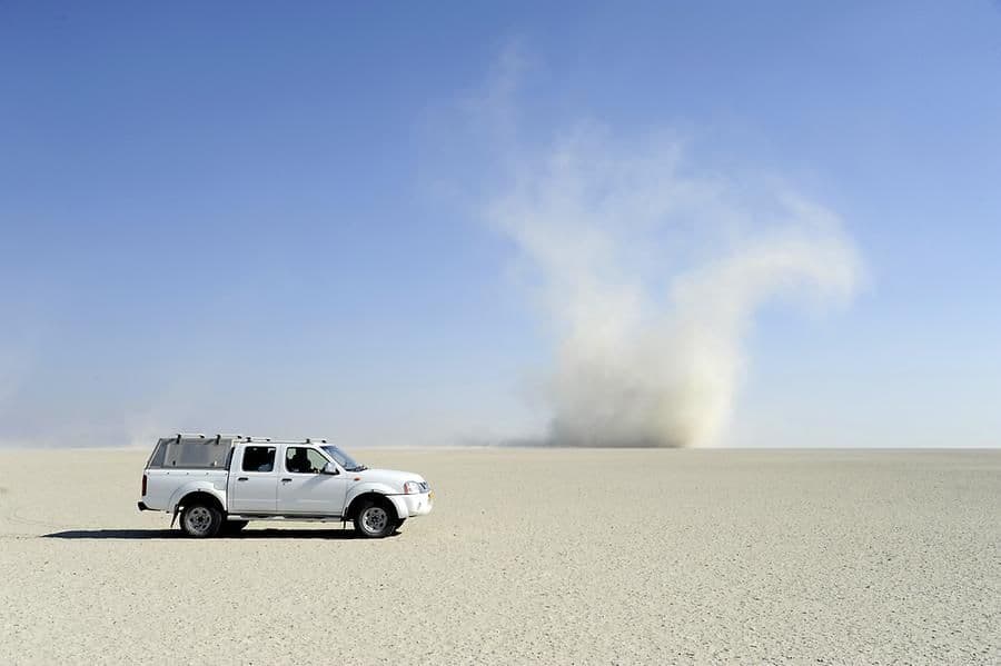 Un pickup bianco è parcheggiato in un vasto deserto piatto mentre un grande diavolo di polvere turbina in lontananza sotto un cielo azzurro chiaro.