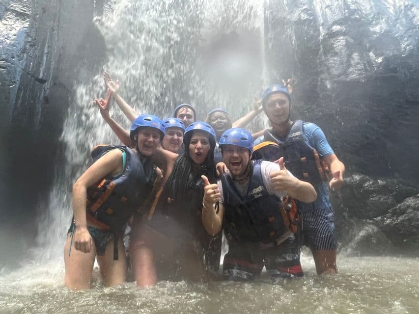 Un groupe WeRoad en voyage, équipé de casques et de gilets de sauvetage, pose pour une photo dans l'eau au pied d'une puissante cascade.