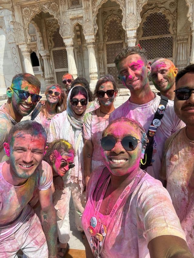 Un groupe WeRoad pose pour un selfie, couvert de poudre colorée Holi devant un bâtiment orné.