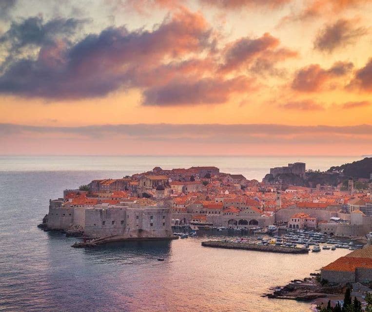 Una vista aérea de una ciudad costera amurallada con tejados de terracota y un puerto lleno de barcos bajo un cielo de atardecer colorido.