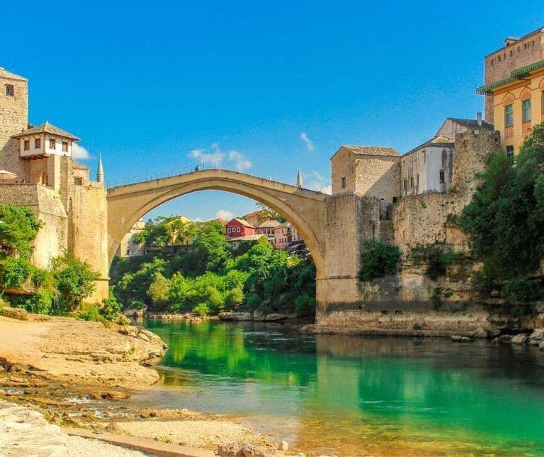 Un puente de piedra arqueado cruza un río turquesa, flanqueado por edificios históricos de piedra y árboles verdes bajo un cielo azul claro.