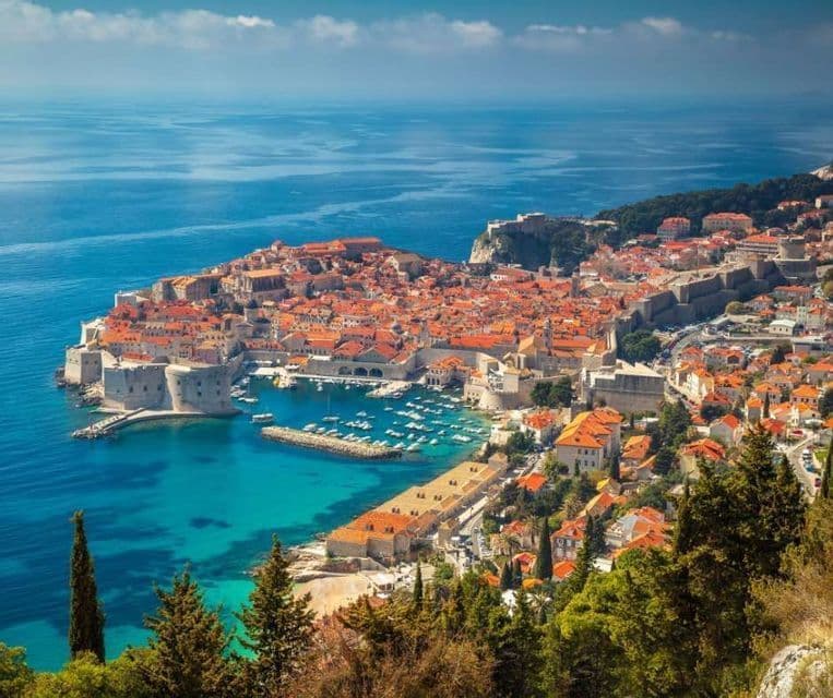 Vista aérea de una ciudad costera amurallada, con tejados de terracota, un pequeño puerto con barcos y el mar azul que la rodea.