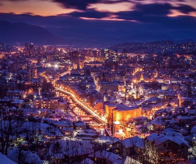 Un paisaje urbano al anochecer con edificios nevados y farolas brillantes, visto desde un punto elevado.