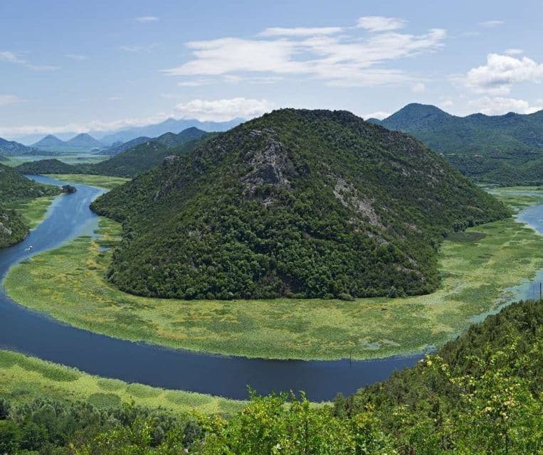 Un ancho río azul forma un meandro en herradura alrededor de una gran colina cubierta de árboles, con más colinas al fondo bajo un cielo parcialmente nublado.