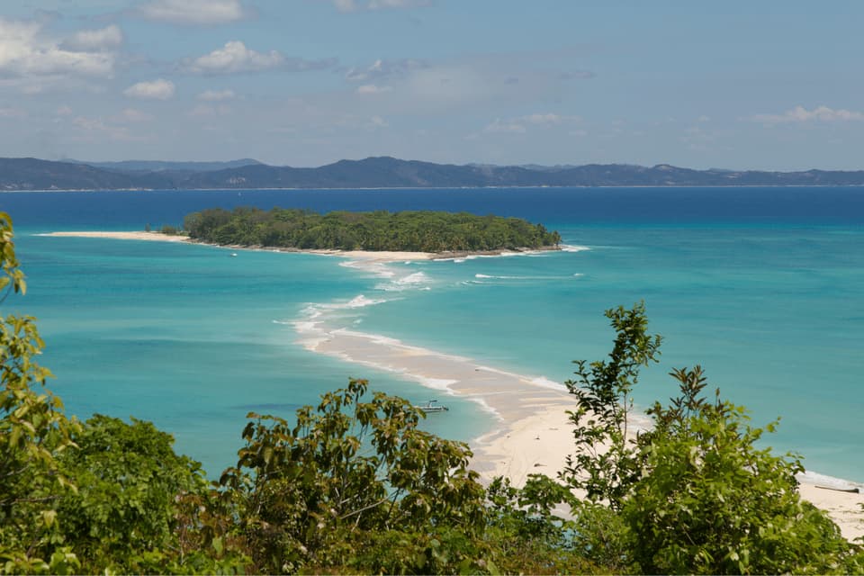 Ein erhöhter Blick auf eine schmale, weiße Sandbank, die mit einer bewaldeten Insel verbunden ist, umgeben von türkisfarbenem Ozeanwasser.