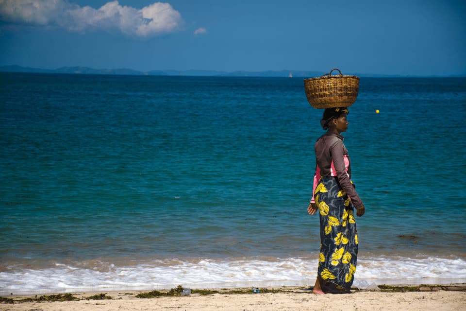 Eine Frau mit einem Weidenkorb auf dem Kopf steht an einem Sandstrand neben dem blauen Meer.