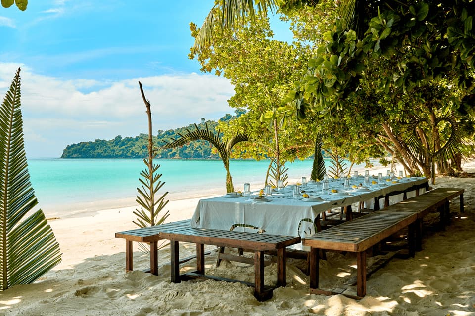 Ein langer Esstisch mit Holzbänken, gedeckt für eine Mahlzeit an einem Sandstrand mit Blick auf einen türkisfarbenen Ozean.