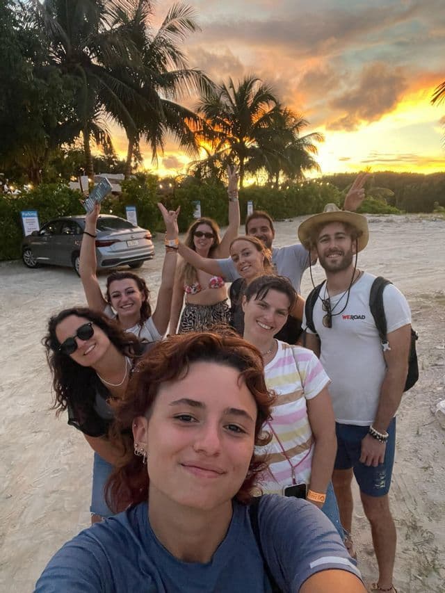 Un viaggio di gruppo WeRoad in posa per un selfie di gruppo su una spiaggia al tramonto, con palme sullo sfondo.