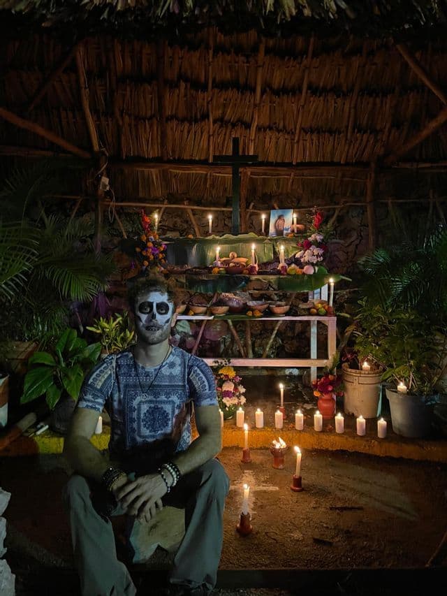 Un uomo con il viso dipinto da teschio siede di fronte a un altare illuminato da candele, decorato con fiori e offerte sotto un tetto di paglia.