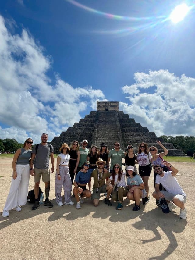 Un gruppo WeRoad in viaggio posa per una foto di gruppo davanti a una grande piramide di pietra sotto un cielo soleggiato e parzialmente nuvoloso.