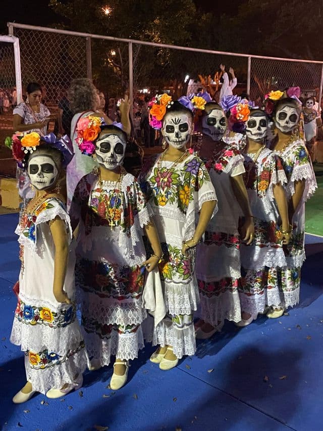 Una fila di donne in abiti tradizionali ricamati e trucco da calavera posano durante un festival notturno.