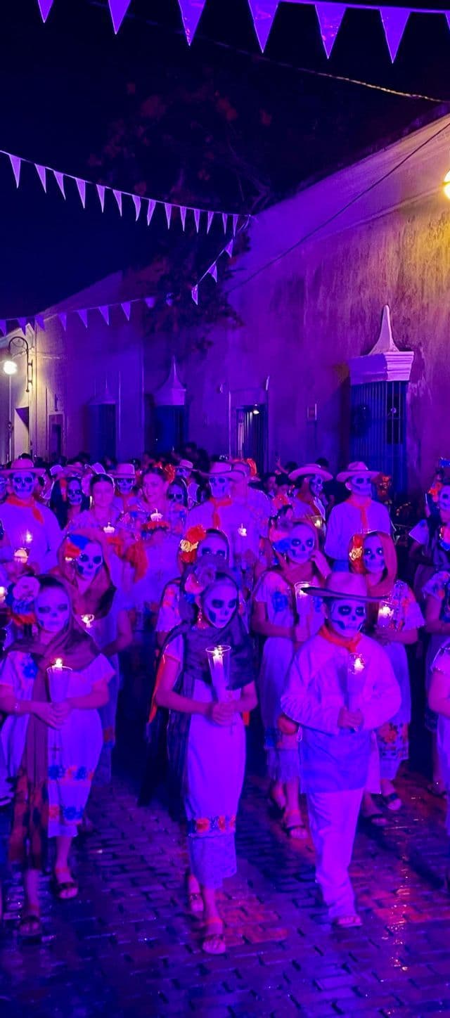 Una folla di persone con trucco da teschio e candele partecipa a una processione notturna su una strada con illuminazione viola.