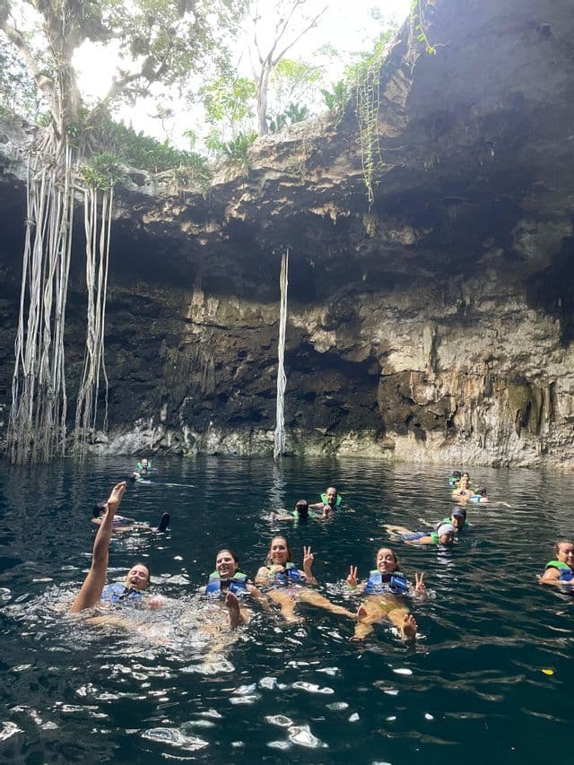 Un gruppo WeRoad nuota in un cenote, circondato da pareti rocciose e radici di alberi pendenti dall'apertura sovrastante.