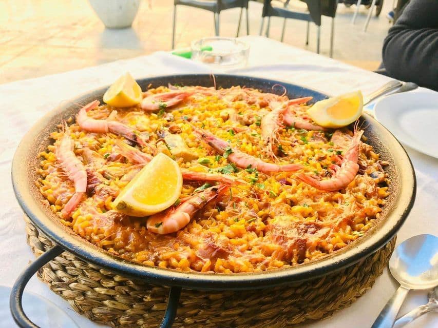 Eine große Pfanne Meeresfrüchte-Paella, garniert mit Garnelen und Zitronenspalten, auf einem geflochtenen Ständer auf einem Tisch.