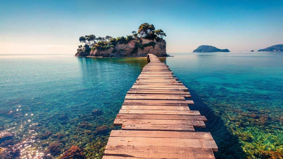 Un paseo de madera se extiende sobre aguas claras de color turquesa hacia una pequeña isla rocosa con árboles bajo un cielo azul brillante.