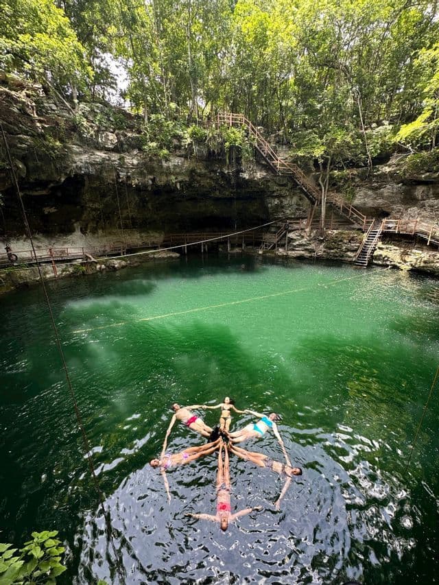 Eine WeRoad-Gruppe bildet eine Sternform, indem sie händchenhaltend im smaragdgrünen Wasser einer Cenote treibt.