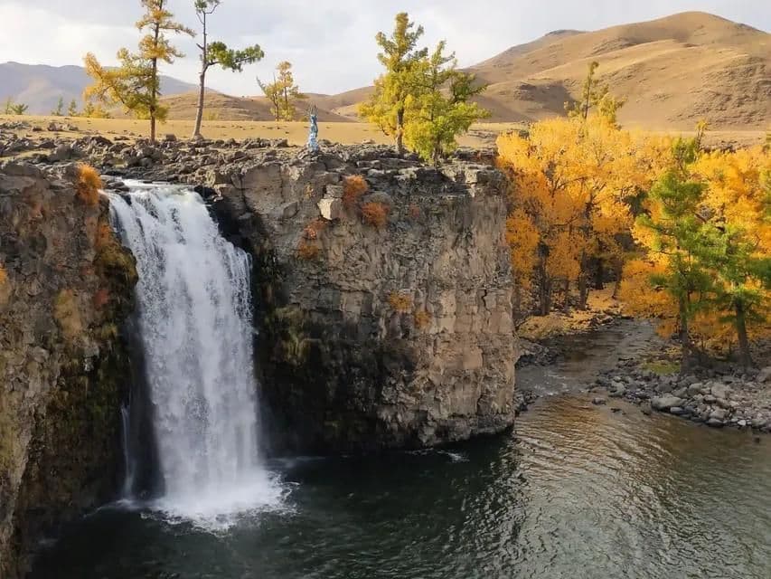 Ein breiter Wasserfall stürzt über eine felsige Klippe in einen Fluss, umgeben von Bäumen mit gelben Herbstblättern und sanften Hügeln im Hintergrund.