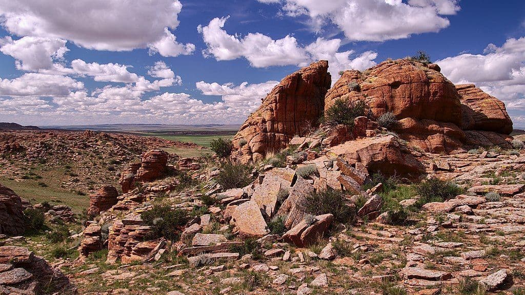 Eine weite Landschaft aus rötlichen Felsformationen und spärlicher Vegetation unter einem strahlend blauen Himmel, gefüllt mit weißen Wolken.