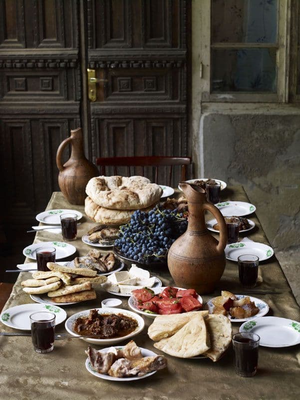 Un tavolo rustico imbandito con un pasto tradizionale a base di focaccia, uva, formaggio, carne e vino in brocche di argilla.