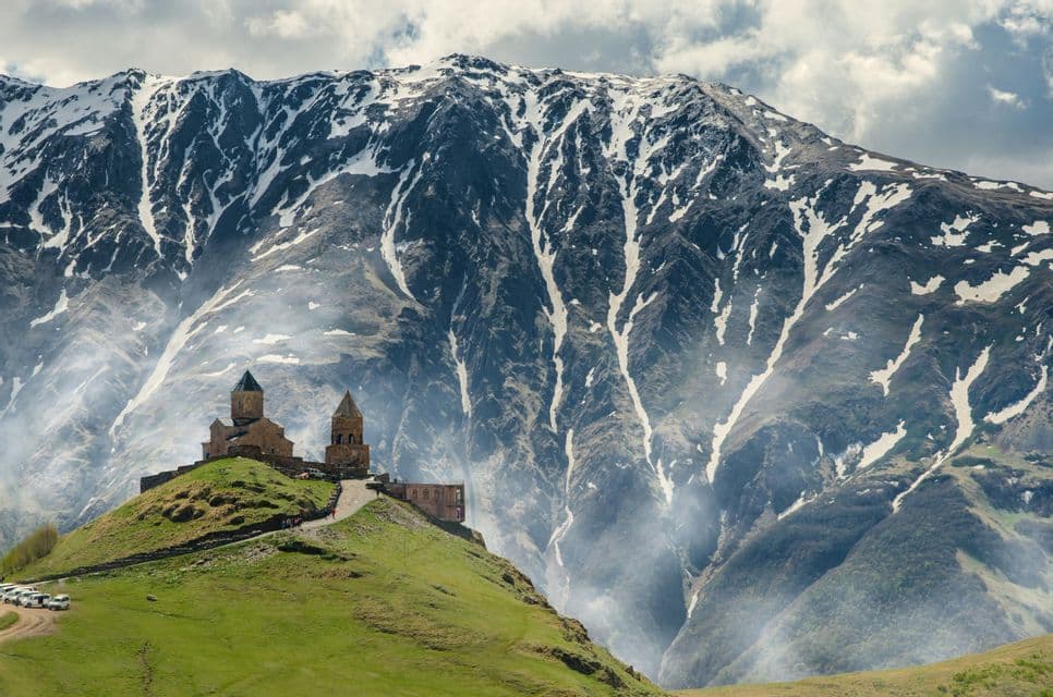 Un'antica chiesa in pietra con torri, su una verde collina con un'imponente montagna striata di neve sullo sfondo.