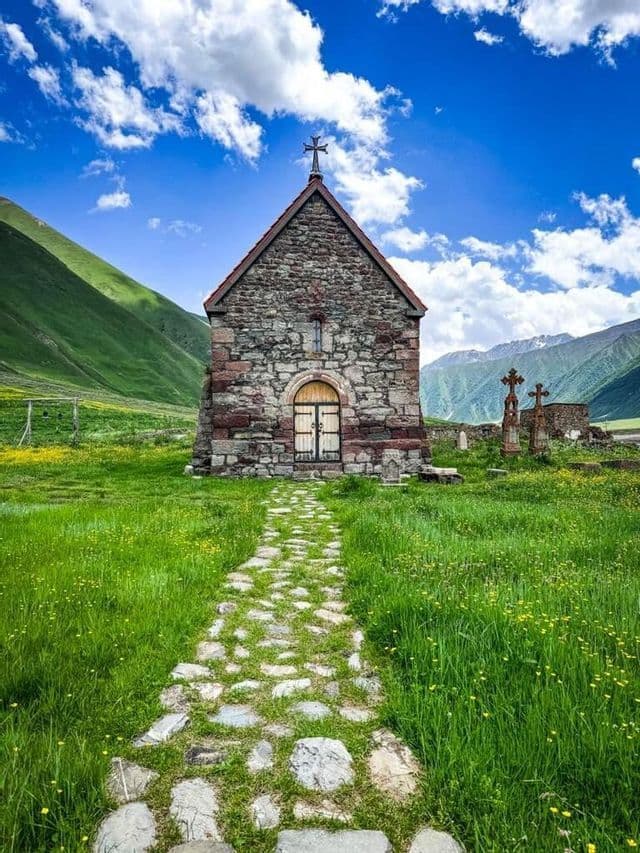 Un sentiero di ciottoli conduce a una storica cappella in pietra in una lussureggiante valle di montagna sotto un cielo parzialmente nuvoloso.