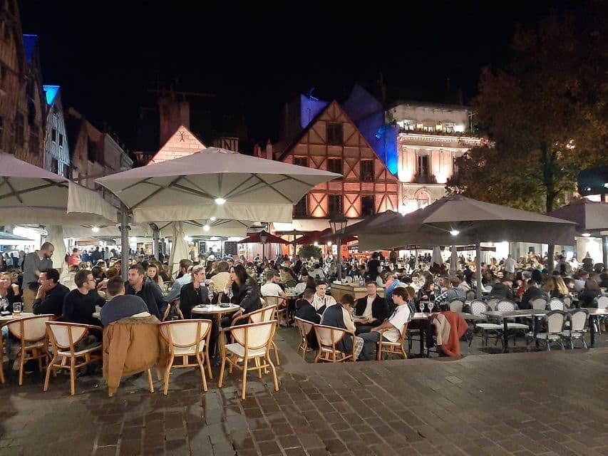 De nombreuses personnes assises à des tables sous de grands parasols dans un café en plein air animé sur une place pavée la nuit.