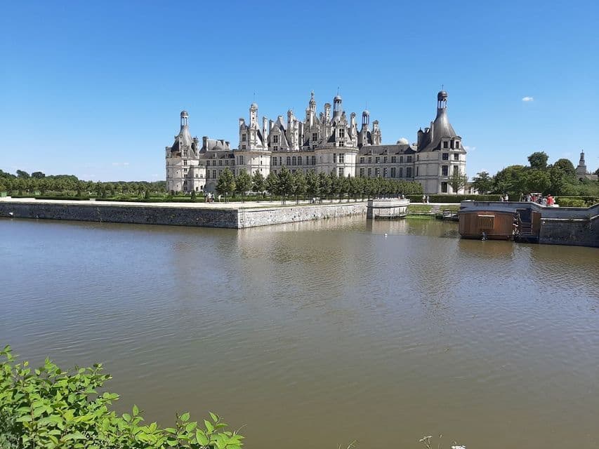 Un château de pierre blanche orné de nombreuses tourelles est vu de l'autre côté de larges douves par une journée claire et ensoleillée.