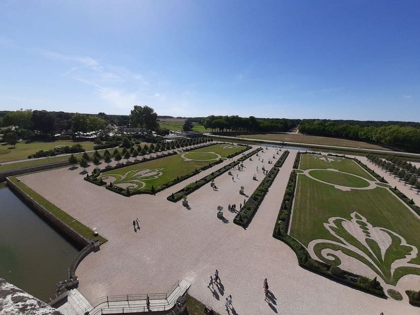 Vue en plongée d'un grand jardin à la française avec des motifs de haies complexes, des pelouses impeccables et des gens marchant sur des allées de gravier.